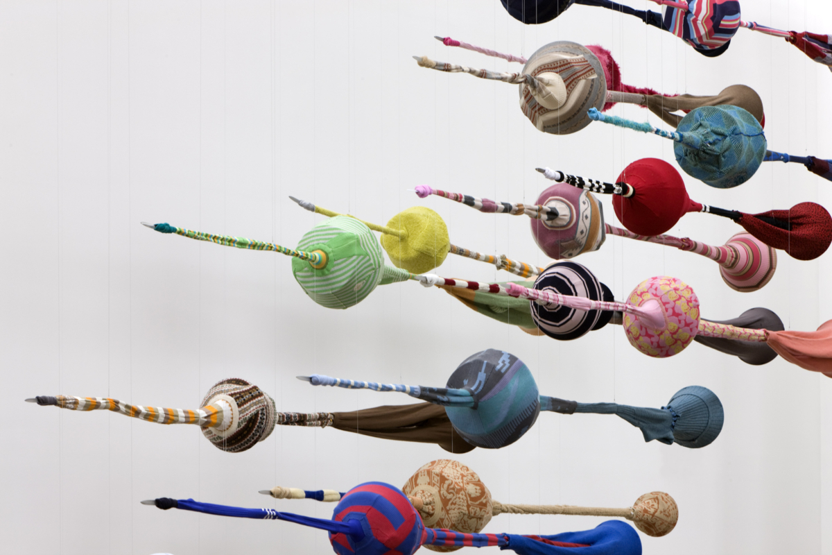 Sculpture of hanging spear-like objects upholstered with used textiles.